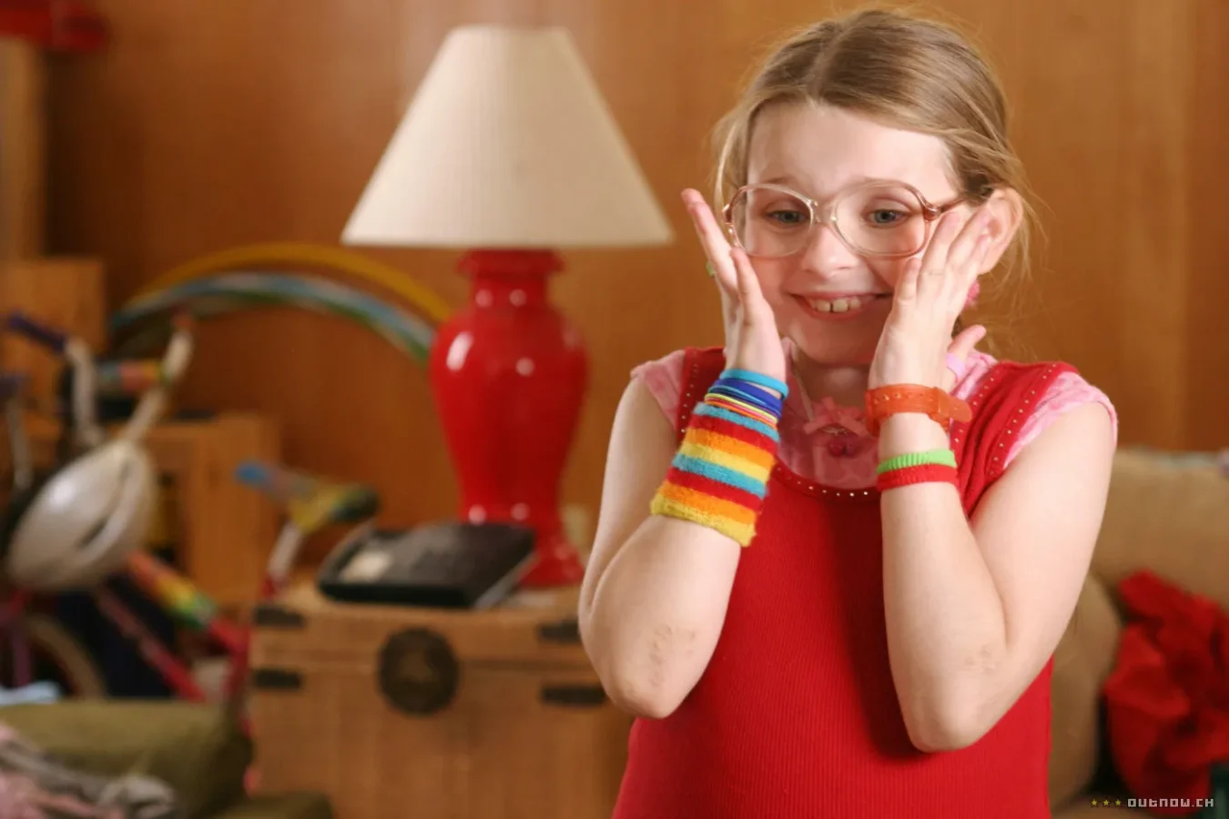 Abigail Breslin, atriz mirim, com roupas coloridas e sorrindo, levando as mãos às bochechas. 