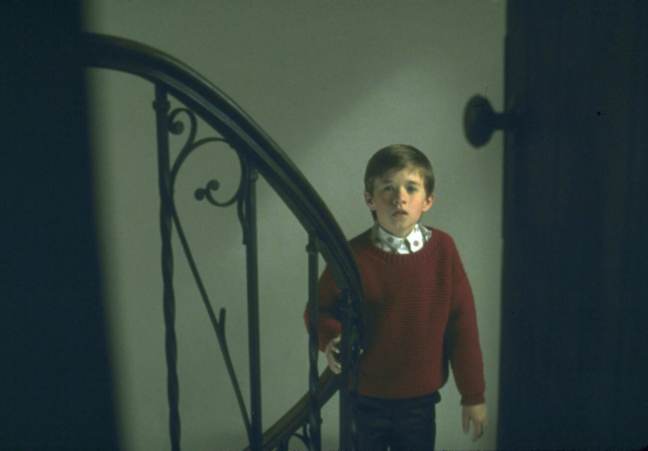 Haley Joel Osment, ator mirim, em filme de terror, ao lado de um corrimão de escada olhando para dentro de uma porta, onde está a câmera. Assim, ele olha para o telespectador.