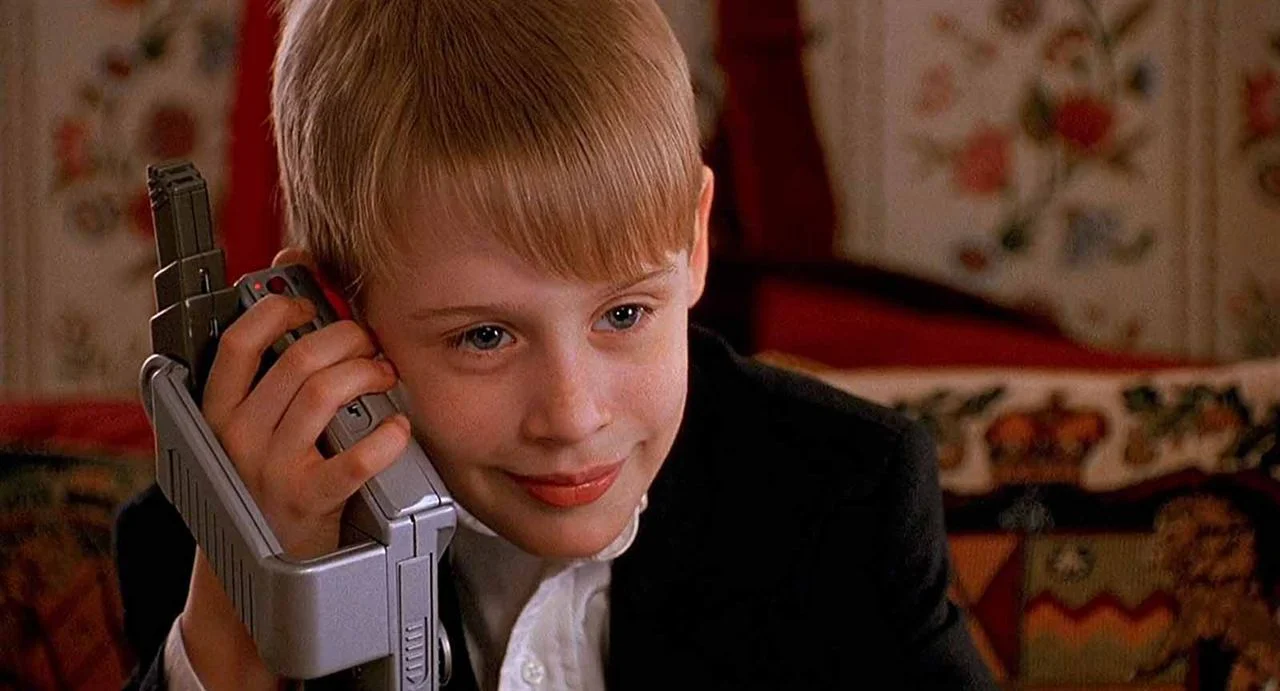 Macaulay Culkin, ator mirim, sorrindo segurando um objeto eletrônico dos anos 80 encostado no ouvido.