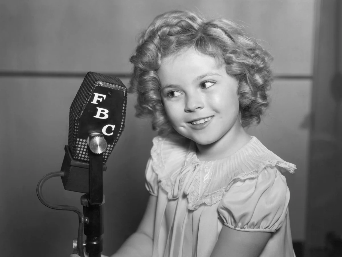 Shirley Temple criança ao lado de um microfone, sorrindo, em foto preto e branco