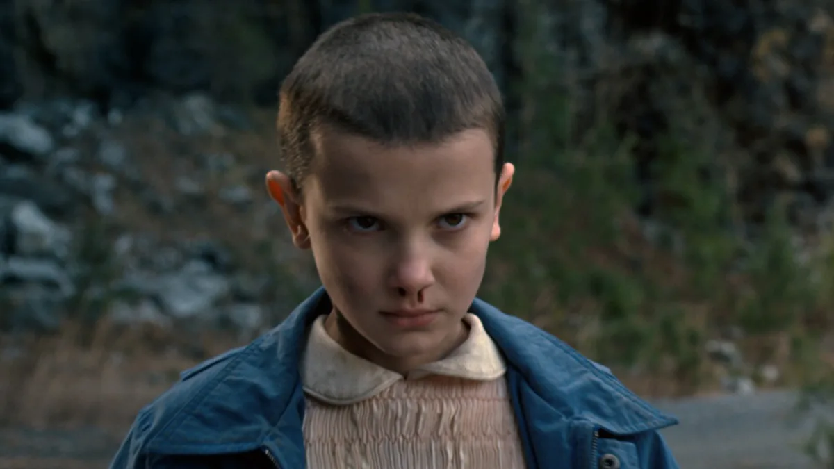 Millie Bobby Brown, atriz mirim, em Stranger Things, com o cabelo raspado, olhar sério e sangue saindo do nariz.