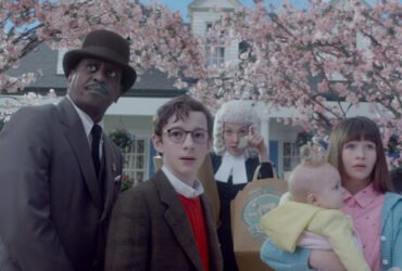 Cinco pessoas reunidas em frente a uma casa com árvores com folhas cor de rosa. Da esquerda para a direita, um homem alto e negro usando terno e chapéu, um menino branco de óculos, uma mulher branca com peruca de juíz branca, uma menina branca segurando um bebê branco no colo. Créditos: Desventuras em Série, Netflix