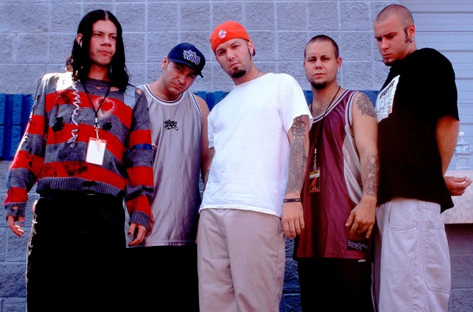 Cinco integrantes da banda Limp Bizkit posam para uma foto casual ao ar livre, com roupas largas e estilos distintos.