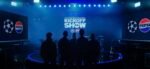 A banda Linkin Park, composta por cinco homens e uma mulher, está parada em frente a um painel de led com cores azuladas escrito "UEFA Champions League Final Kick-off show"