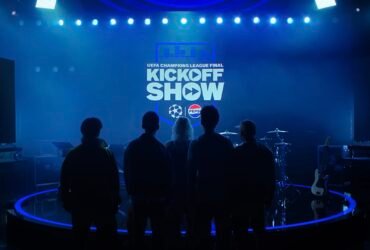 A banda Linkin Park, composta por cinco homens e uma mulher, está parada em frente a um painel de led com cores azuladas escrito "UEFA Champions League Final Kick-off show"