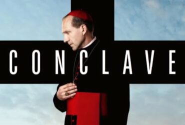 O pôster do filme Conclave, tem o personagem Cardeal Lawrence vestindo as roupas tradicionais de Cardeal nas cores preto e vermelho. O fundo da imagem é um céu azul e na frente em destaque está o título do filme.