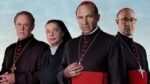 Quatro personagens do filme conclave posam para a câmera. Três deles são homens e uma é uma mulher. Os homens são cardeais candidatados ao cargo de Papa e a mulher é uma freira. Todos os indivíduos são brancos.