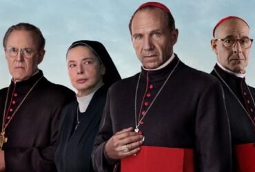 Quatro personagens do filme conclave posam para a câmera. Três deles são homens e uma é uma mulher. Os homens são cardeais candidatados ao cargo de Papa e a mulher é uma freira. Todos os indivíduos são brancos.