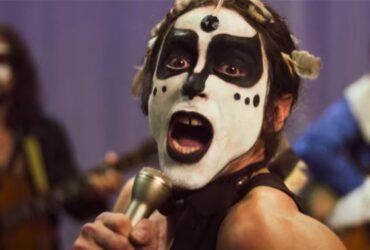 A imagem é uma cena do filme Homem com H. Em primeiro plano está o ator Jesuíta Barbosa caracterizado como Ney Matogrosso uma pessoa do gênero masculino segurando um microfone e cantando com a boca aberta mostrando os dentes. Essa pessoa usa uma maquiagem branca no rosto mas seus olhos e sua boca estão cobertos por maquiagem preta. Ao fundo há duas pessoas também do sexo masculino tocando violão. O da direita está cantando em um microfone preso a um pedestal.