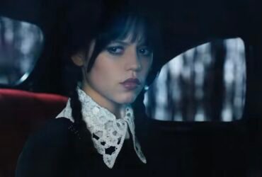 A personagem Wandinha está sentada no banco de trás de um carro preto. Ela olha pela janela com uma expressão de desconfiança.