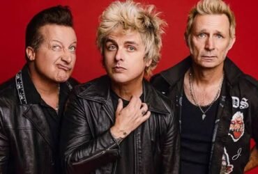 Os três integrantes da banda Green Day posam para foto de divulgação. Os três são homens brancos de olhos claros e vestem roupas pretas.