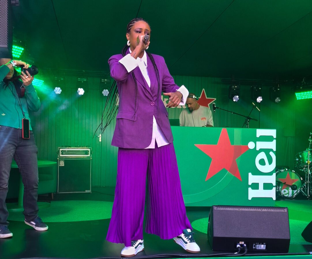 A rapper Stefanie é uma mulher negra, ela usa traças no cabelo, uma calça larga e um terno, ambos na cor roxa, enquanto canta em um palco na cor verde.