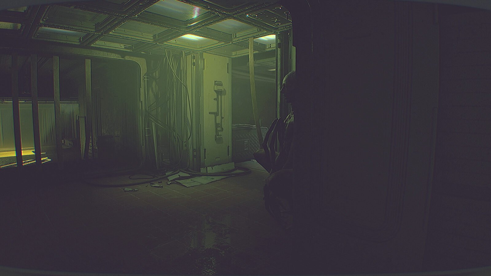 Interior de uma instalação escura e abandonada com iluminação verde-amarelada fraca no teto. Cabos elétricos expostos e detritos espalhados pelo chão de ladrilhos molhados. Uma figura parcialmente visível se esconde atrás de um painel escuro à direita, observando o ambiente.