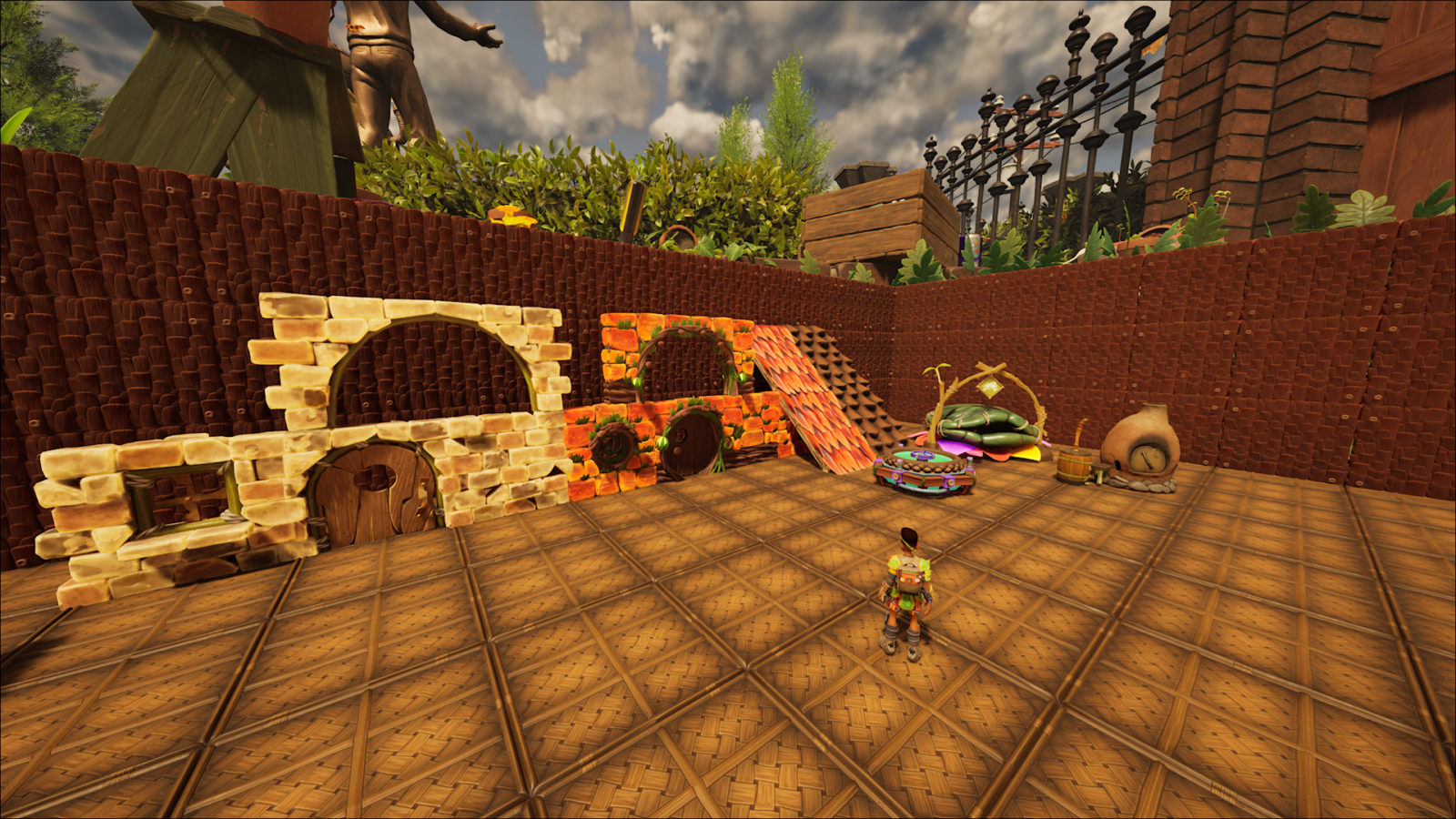 Captura de tela do jogo Grounded mostrando um personagem em um amplo pátio de palha trançada. Ao fundo, uma parede de madeira exibe diferentes estilos de construção: estruturas de pedra com arcos, paredes de cogumelos alaranjados, uma rampa de pétalas e estações de trabalho como um forno de argila e uma cama de pétalas.