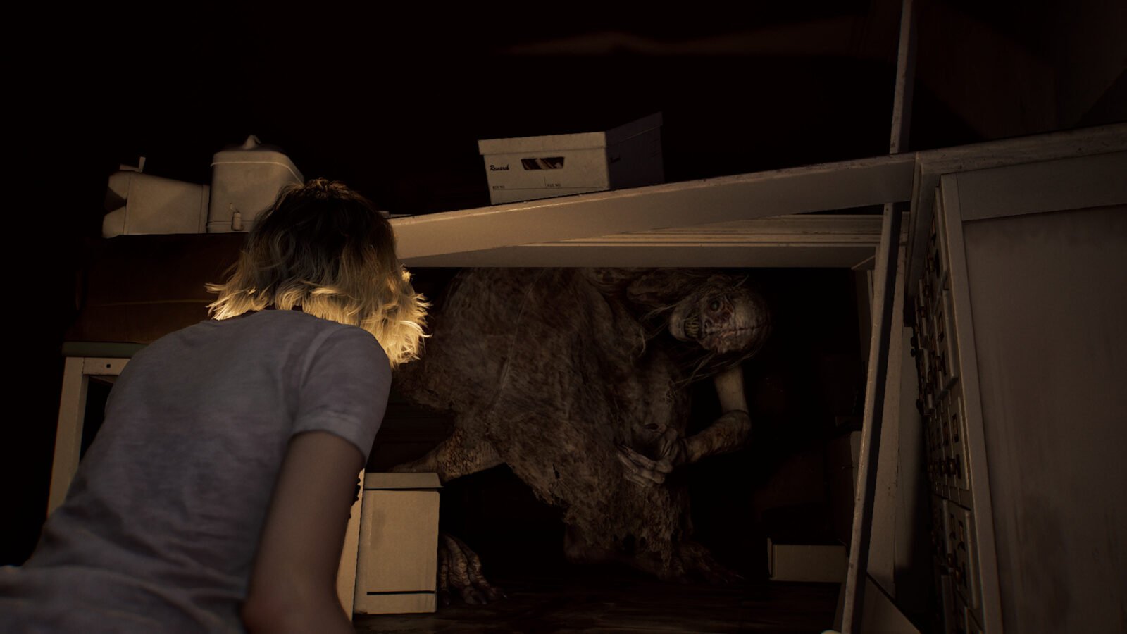 Close-up por trás de um Grace, agachada no chão. Ela está voltada para a direita, olhando para a Garota, um grande e horrendo monstro sob uma mesa de escritório. O monstro tem pele pálida e deformada e olhos grandes e escuros que olham para a personagem. O ambiente é escuro e bagunçado, com caixas de papelão e outros objetos de escritório espalhados. Apenas a parte superior da personagem e o monstro sob a mesa estão visíveis, com o resto da sala mergulhado em sombras densas.