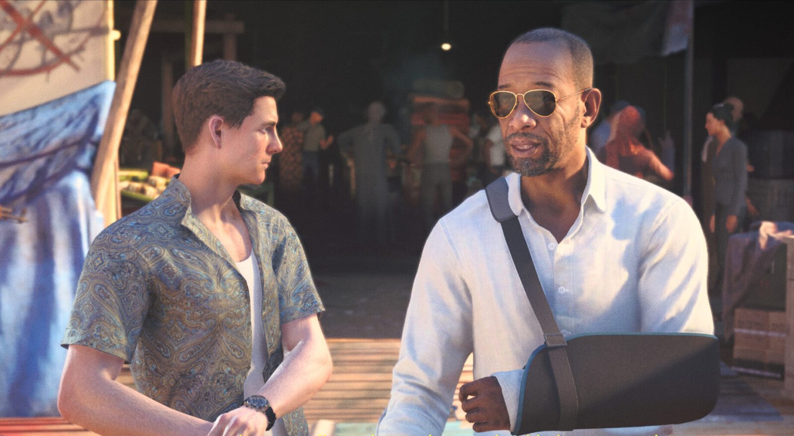 Captura de tela de uma cena de First Light. À esquerda, um jovem James Bond, de perfil, usa uma camisa curta estampada em tons de azul e dourado sobre uma camiseta branca, olhando para o lado. À direita, John Greenway, com barba por fazer, óculos de sol estilo aviador e camisa social branca, está com o braço esquerdo imobilizado em uma tipoia escura. O fundo mostra um ambiente externo movimentado e sombreado com outras pessoas borradas.