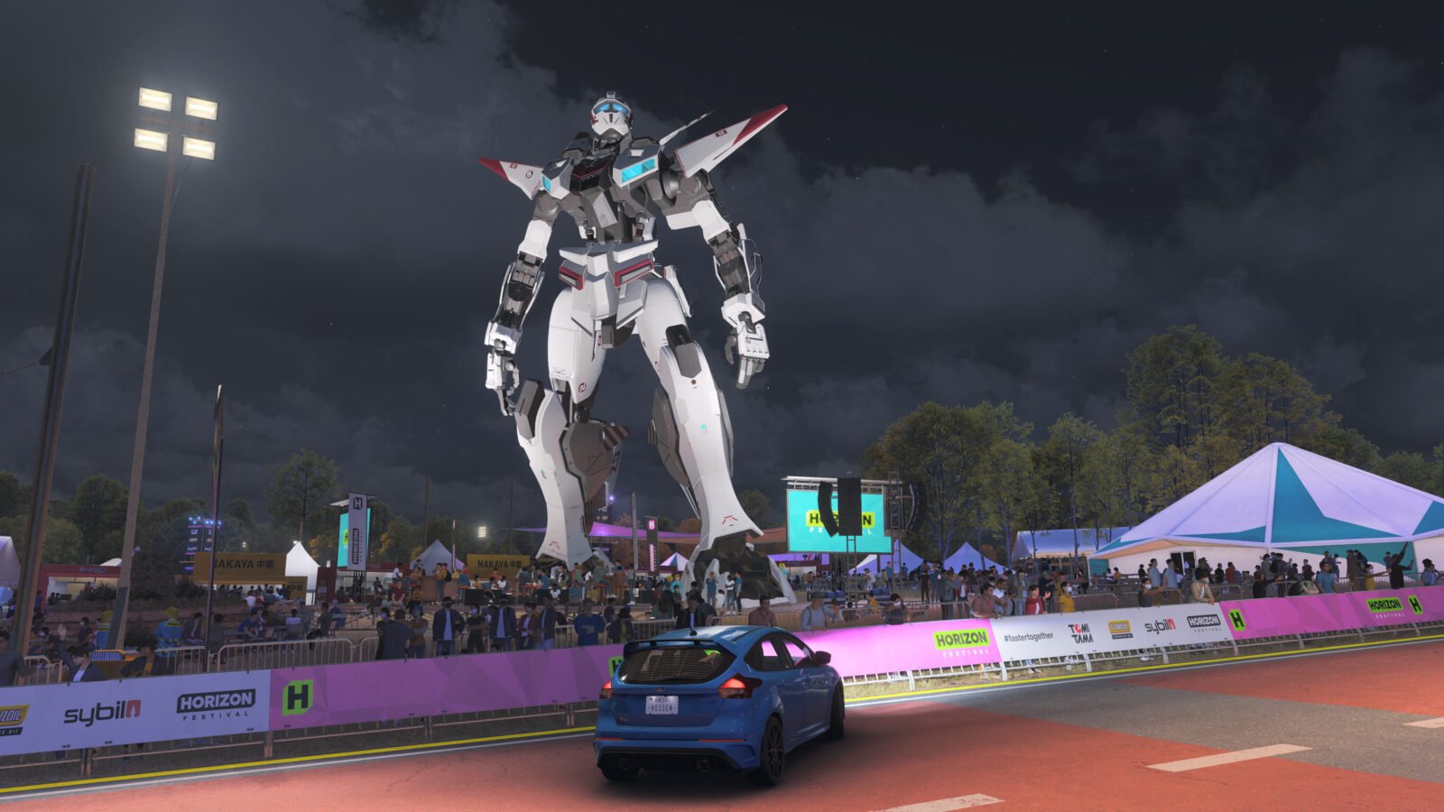 Um carro azul parado em frente a um enorme robô futurista branco, posicionado no centro de um festival noturno iluminado com cercas cor-de-rosa e público ao fundo.