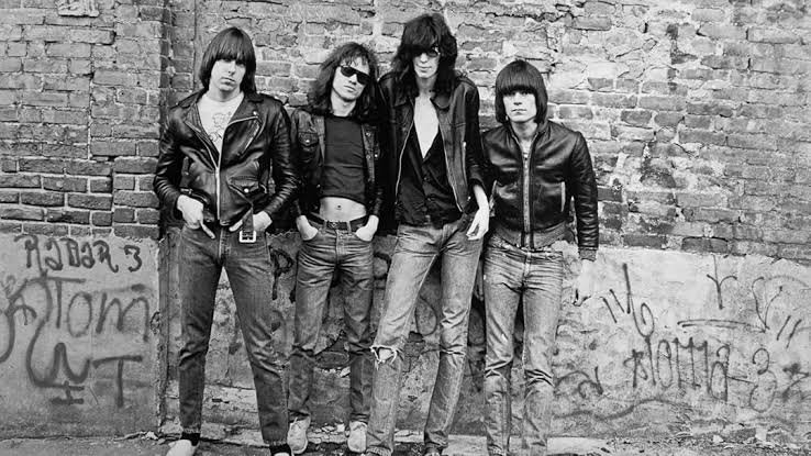 A banda Ramones, formada por quatro homens vestindo jaquetas de couro pretas, posa para foto em frente a uma parede de tijolos. A foto é em preto e branco.