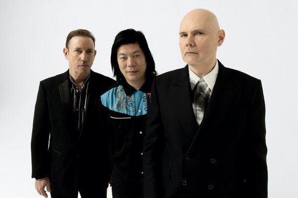 A banda The Smashing Pumpkins, composta atualmente por um trio de homens vestindo roupas pretas posa para foto em um estúdio branco.