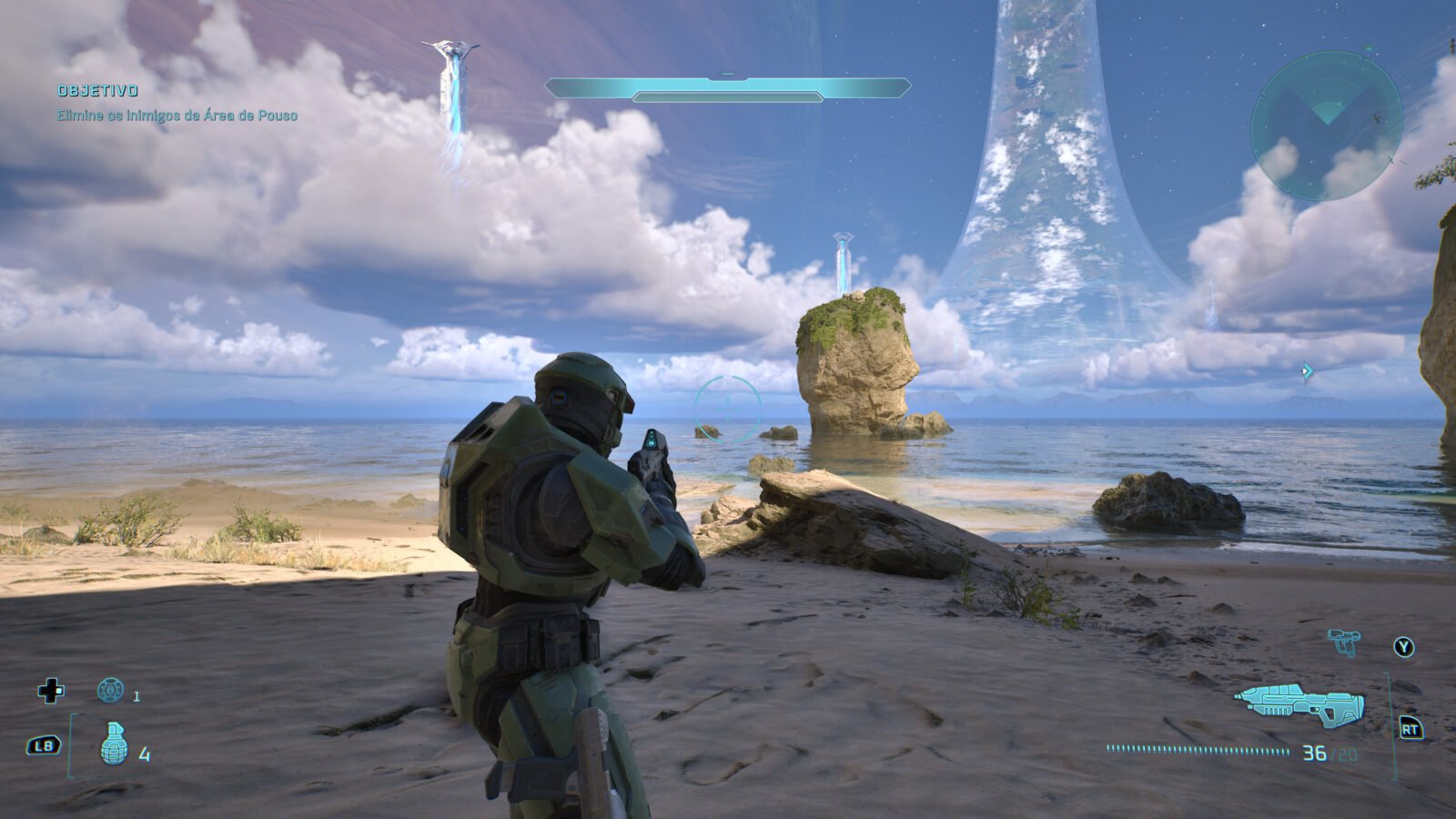 Master Chief, com sua icônica armadura verde, de pé em uma praia arenosa. Ele olha para o mar, onde grandes monólitos de pedra e imponentes pilares de luz alienígenas Forerunner se estendem sob um céu dramático. No horizonte elevado, a gigantesca instalação Halo curva-se em direção às nuvens e um planeta gigante gasoso. O HUD do jogo é visível, incluindo o objetivo em português: 'Elimine os inimigos da Área de Pouso'.