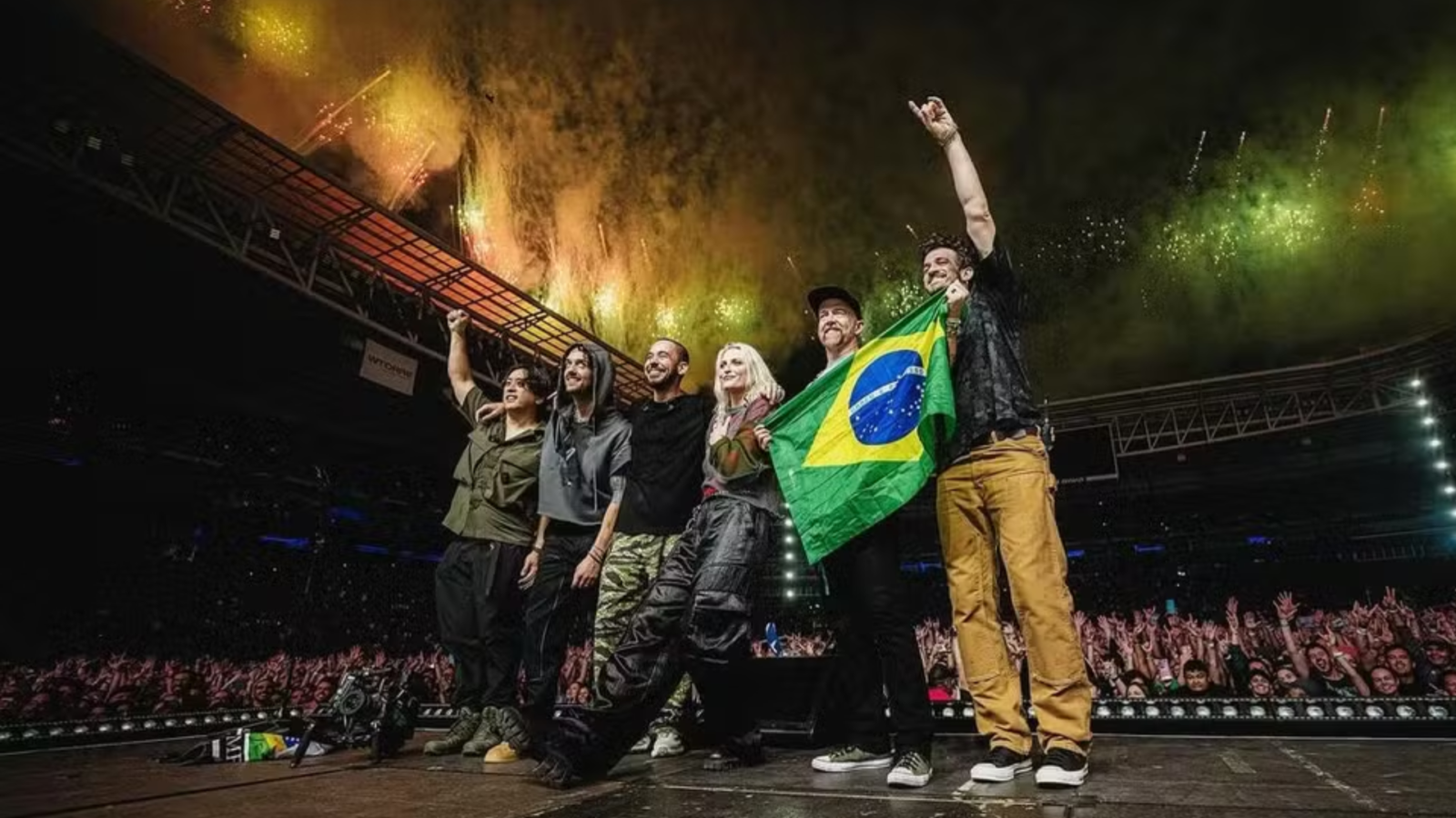 ​Foto do Linkin Park no palco do Allianz Parque em 2024. A banda posa com a bandeira do Brasil diante de uma multidão e fogos de artifício.
