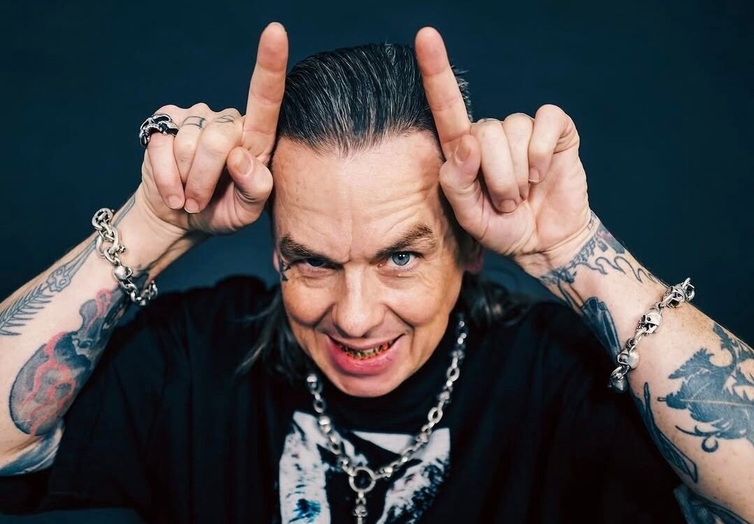 Sid Wilson, um homem de cabelos grisalhos penteados para trás e braços bastante tatuados faz o gesto de chifres apontando os indicadores para o alto da cabeça. Ele sorri de forma provocativa, exibindo dentes com detalhes metálicos, e usa pulseiras e colar de correntes com caveiras sobre uma camiseta preta.