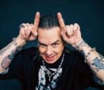 Sid Wilson, um homem de cabelos grisalhos penteados para trás e braços bastante tatuados faz o gesto de chifres apontando os indicadores para o alto da cabeça. Ele sorri de forma provocativa, exibindo dentes com detalhes metálicos, e usa pulseiras e colar de correntes com caveiras sobre uma camiseta preta.