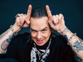 Sid Wilson, um homem de cabelos grisalhos penteados para trás e braços bastante tatuados faz o gesto de chifres apontando os indicadores para o alto da cabeça. Ele sorri de forma provocativa, exibindo dentes com detalhes metálicos, e usa pulseiras e colar de correntes com caveiras sobre uma camiseta preta.