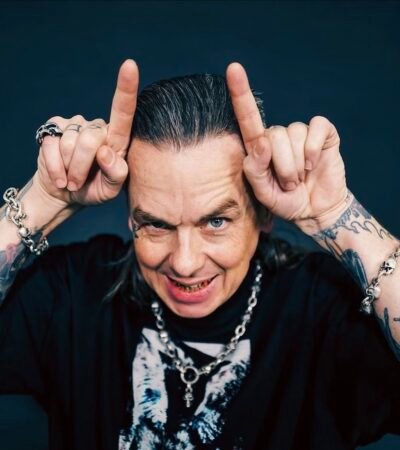 Sid Wilson, um homem de cabelos grisalhos penteados para trás e braços bastante tatuados faz o gesto de chifres apontando os indicadores para o alto da cabeça. Ele sorri de forma provocativa, exibindo dentes com detalhes metálicos, e usa pulseiras e colar de correntes com caveiras sobre uma camiseta preta.