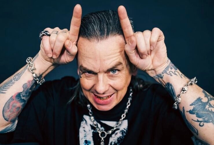 Sid Wilson, um homem de cabelos grisalhos penteados para trás e braços bastante tatuados faz o gesto de chifres apontando os indicadores para o alto da cabeça. Ele sorri de forma provocativa, exibindo dentes com detalhes metálicos, e usa pulseiras e colar de correntes com caveiras sobre uma camiseta preta.