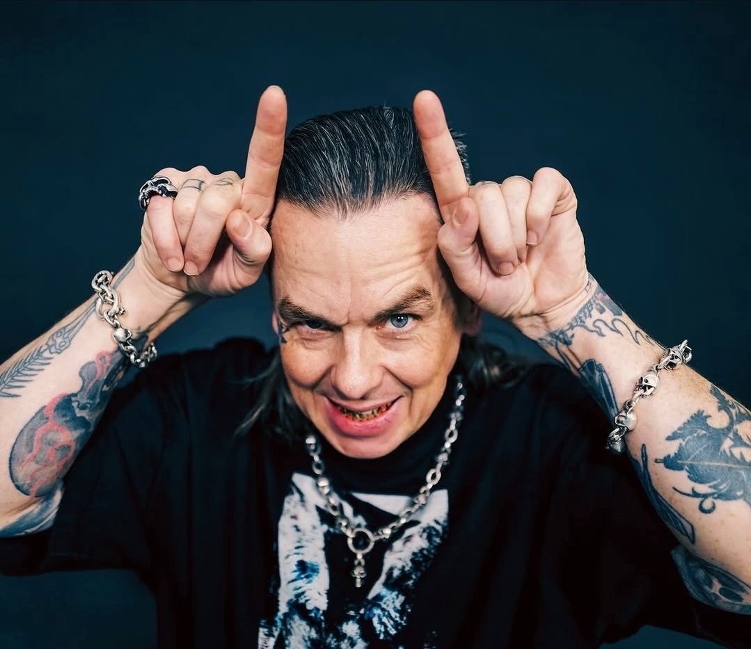 Sid Wilson, um homem de cabelos grisalhos penteados para trás e braços bastante tatuados faz o gesto de chifres apontando os indicadores para o alto da cabeça. Ele sorri de forma provocativa, exibindo dentes com detalhes metálicos, e usa pulseiras e colar de correntes com caveiras sobre uma camiseta preta.